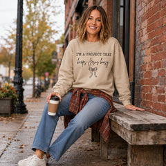 Tea-Shirt Shoppe sand crewneck sweatshirt with I'm a Project That Keeps Jesus Busy text and bow graphic on a woman