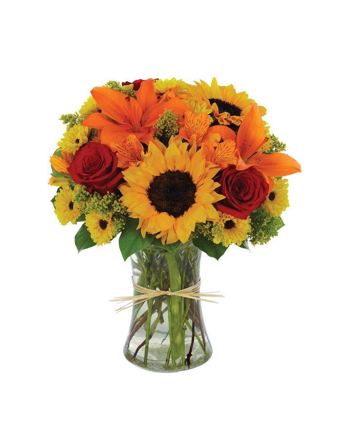 Bouquet of sunflowers, orange lilies, and red roses in a clear vase on a white background
